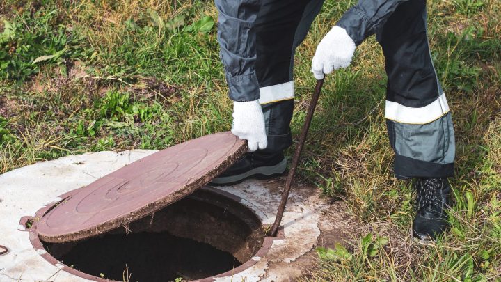 Worker lifting the round manhole cover off a septic tank riser in a yard