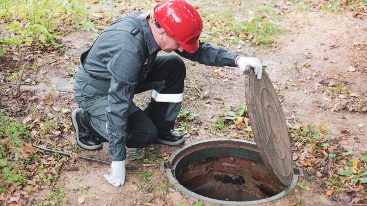 Man in overalls crouched over an open septic tank hatch, looking inside